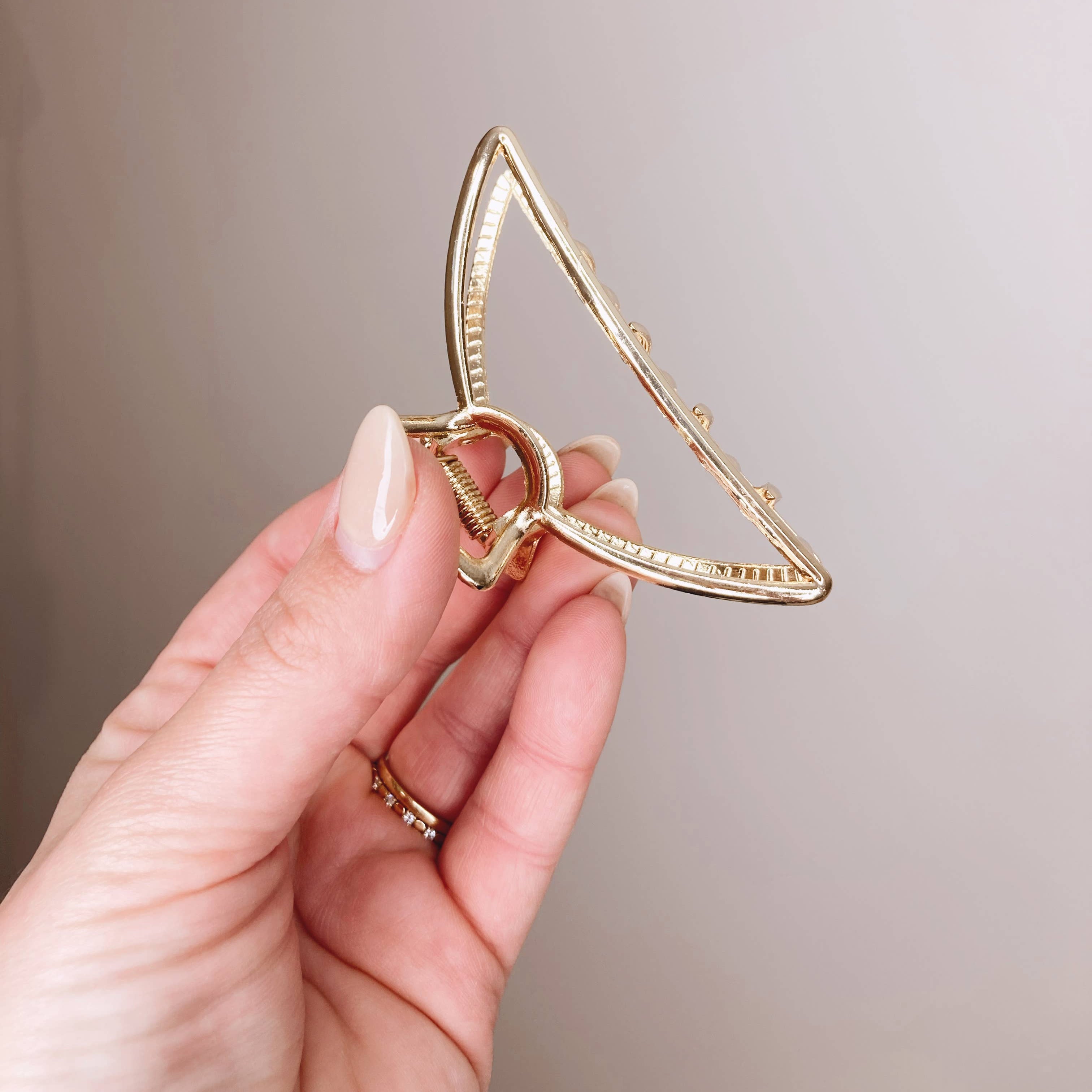 A gold triangular open-frame hair claw clip held by a hand with manicured nails, set against a soft beige background.