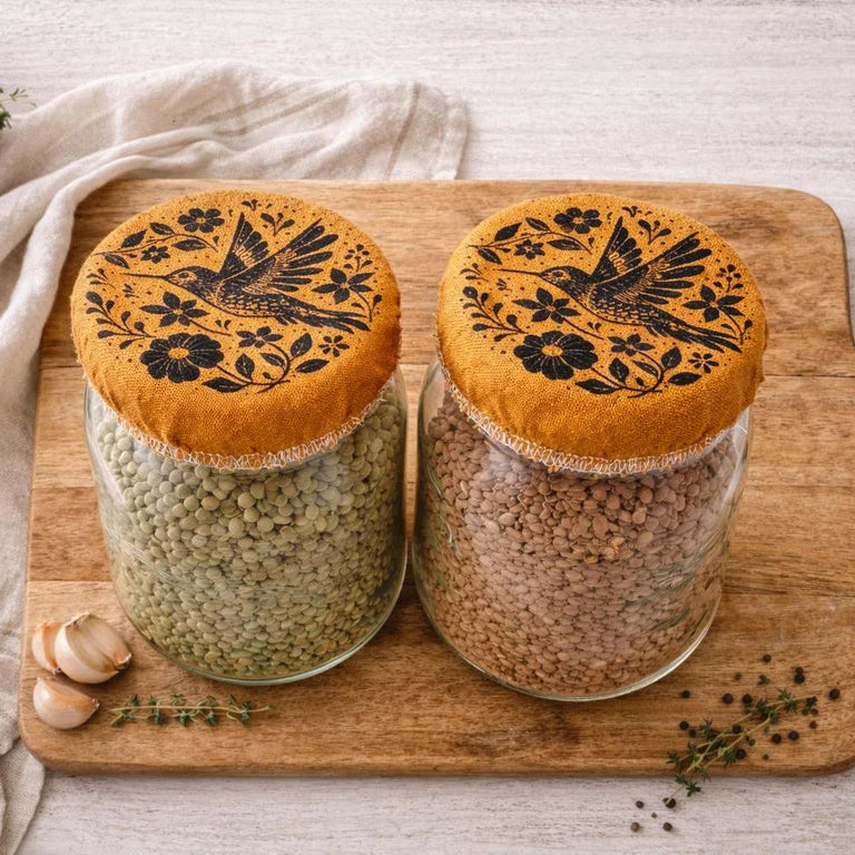 Two glass jars with decorative Golden cloth colored covers with hummingbird design. Placed on a wooden cutting board with herbs and a towel.