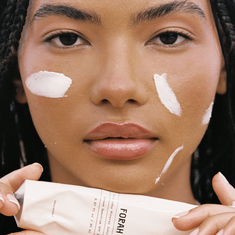 Woman holding a tube of FORAH skincare product with white cream on her face