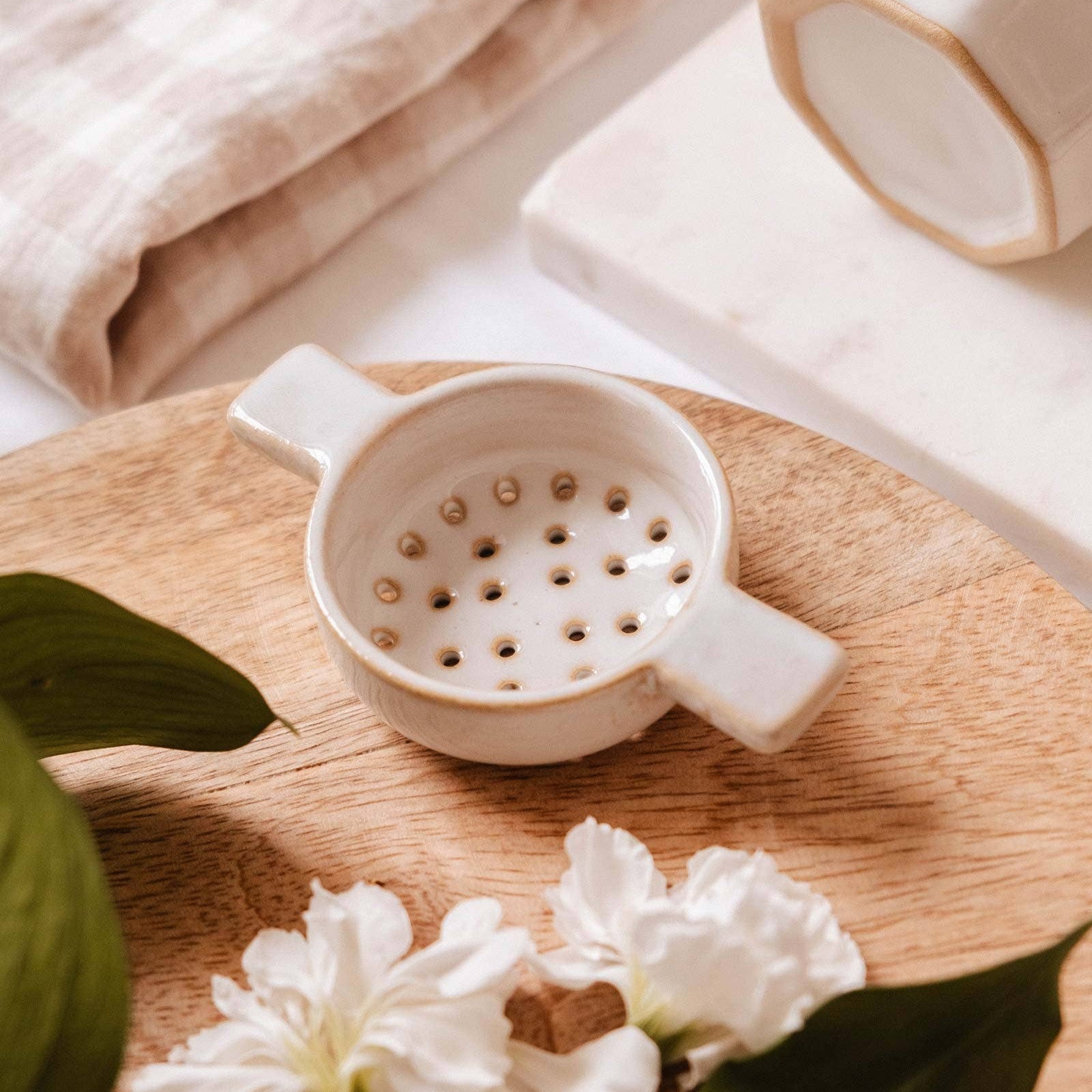 White ceramic strainer on a wooden surface with white flowers and green leaves.