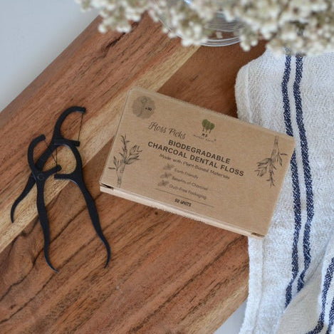A pack of charcoal dental floss picks on a wooden surface, with a white and blue striped cloth and a vase with flowers in the background.