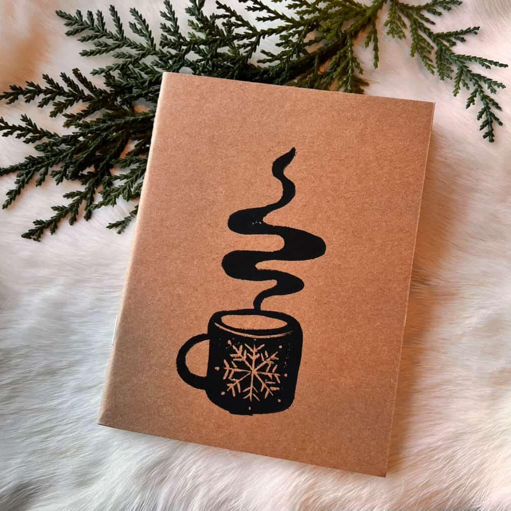 Notebook with steamy coffee mug design on a textured kraft paper surface with greenery and fur background.