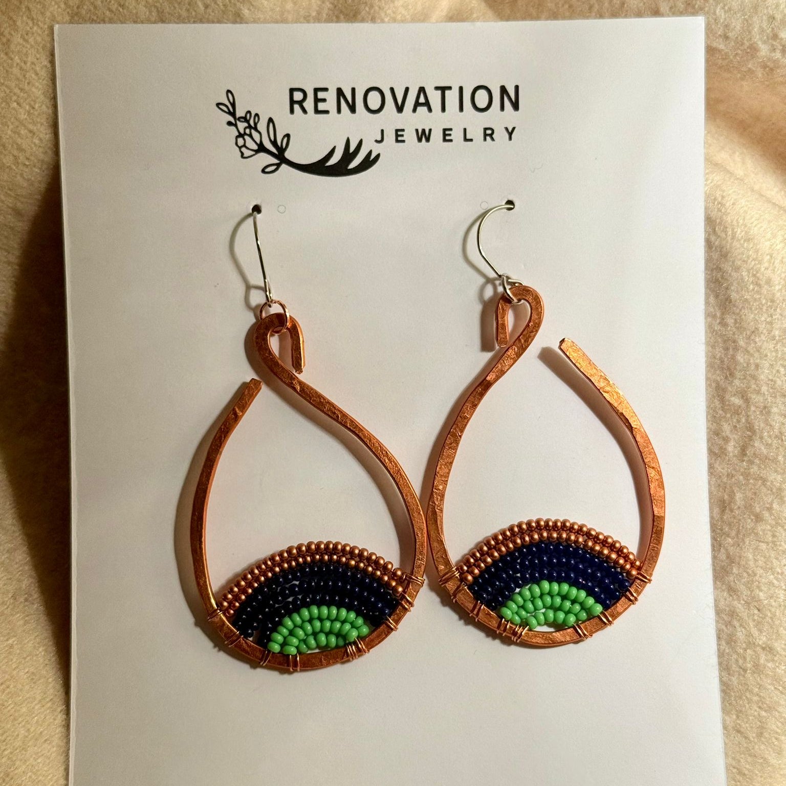 Pair of repurposed copper open hoop earrings on a Renovation Jewelry card with a beige background. Earrings have beads inside the hoops of green, black and brown representing the earth.