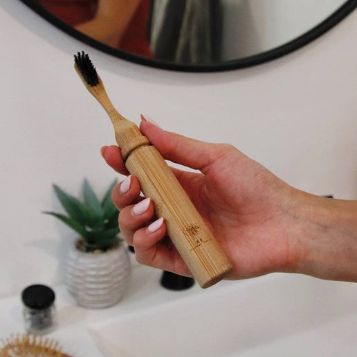 A person holding a bamboo travel toothbrush with a case, placed in a bathroom setting with a mirror, plant, and towel in the background.