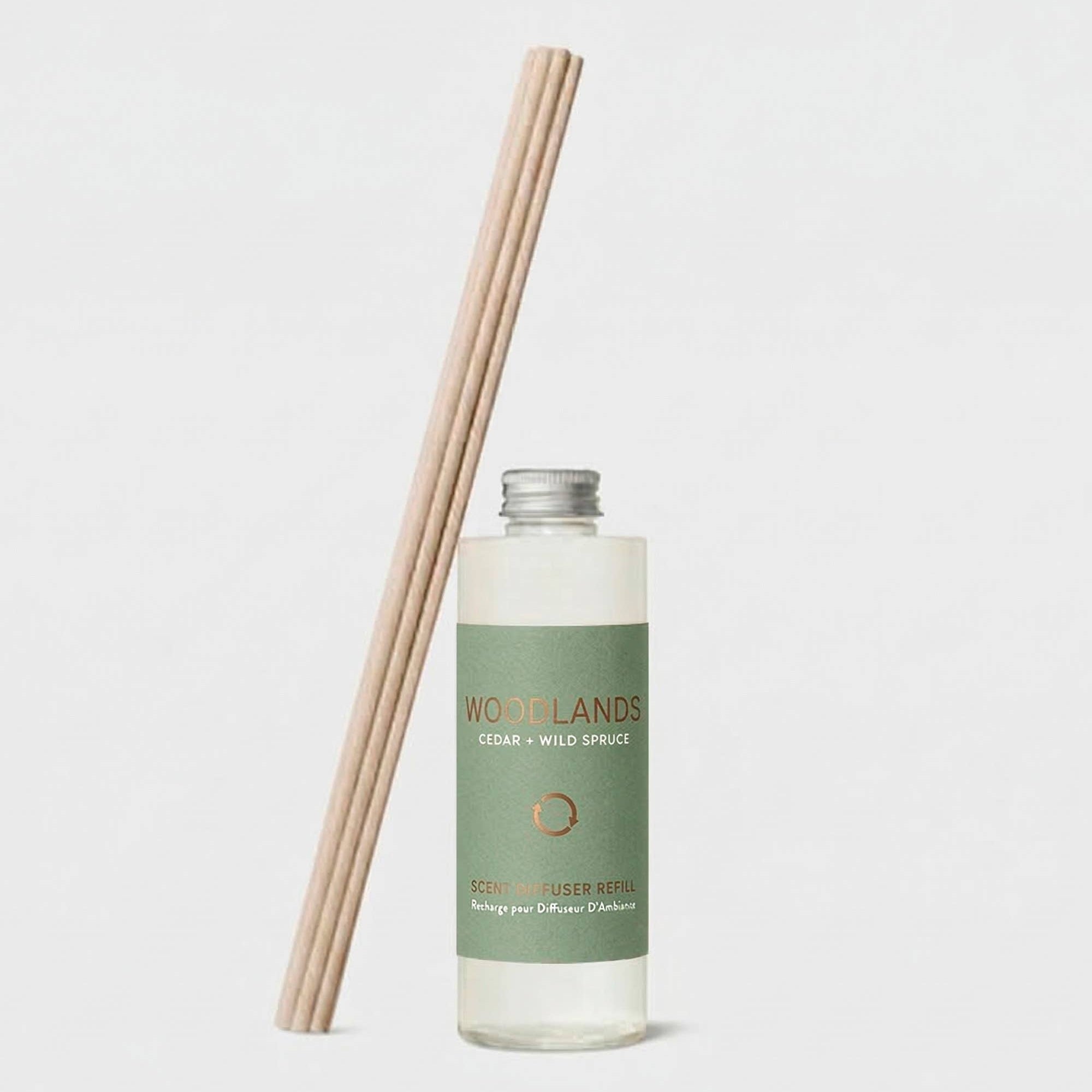 A bottle of Woodlands Cedar + Wild Spruce reed diffuser liquid with a sage green label, shown next to a bundle of natural wood reeds on a light gray background.