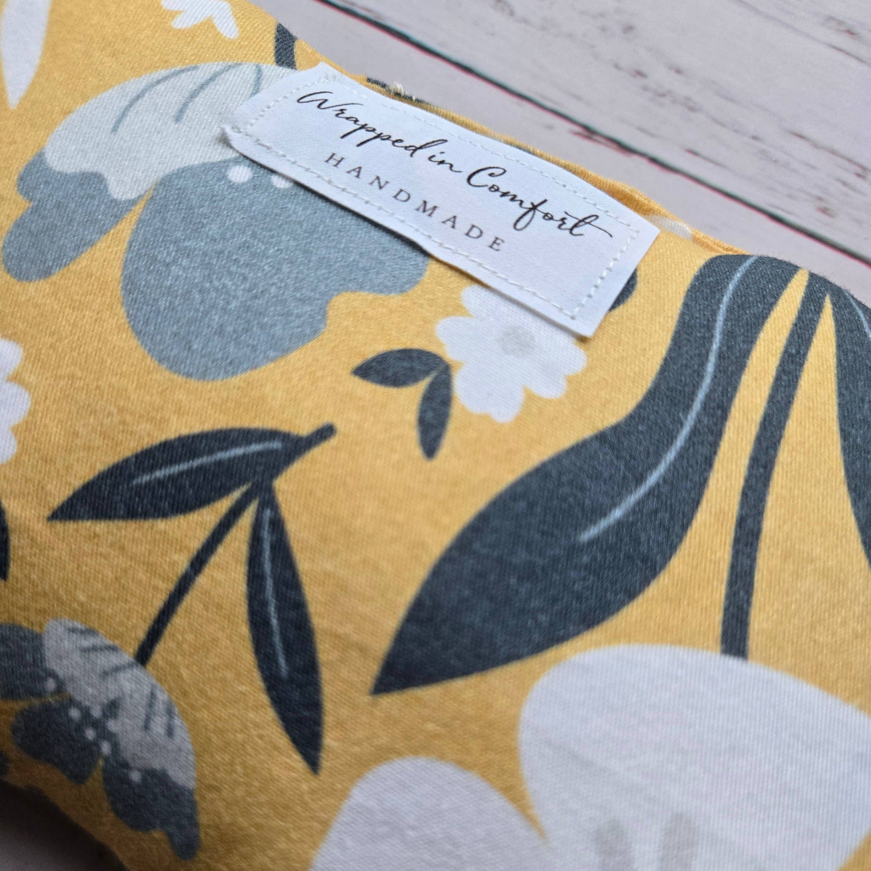 organic cotton bean shaped heating pad with floral-patterned fabric with a 'Wrapped in Comfort' label on a wooden surface