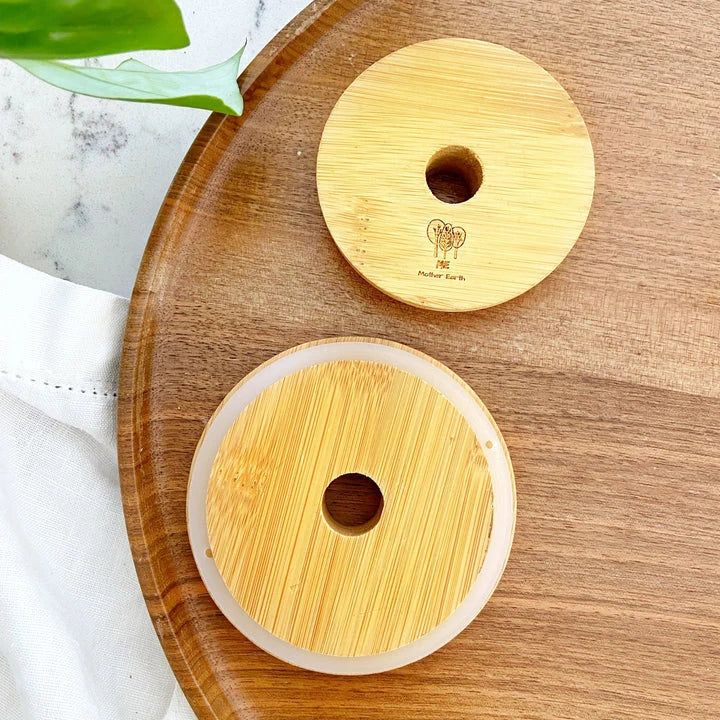 Two round bamboo mason jar covers with a hole for a straw. Has a silicone seal to keep it in place. Engraved with the Me Mother Earth logo, displayed on a wooden tray.
