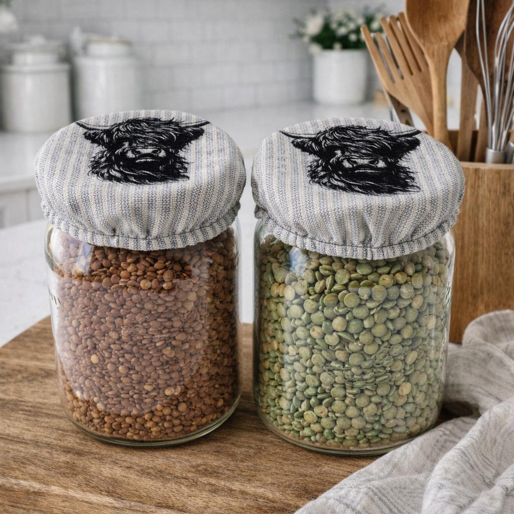 Two jars of dried beans on a wooden cutting board in a kitchen covered with a cloth cover with a highland cow design.