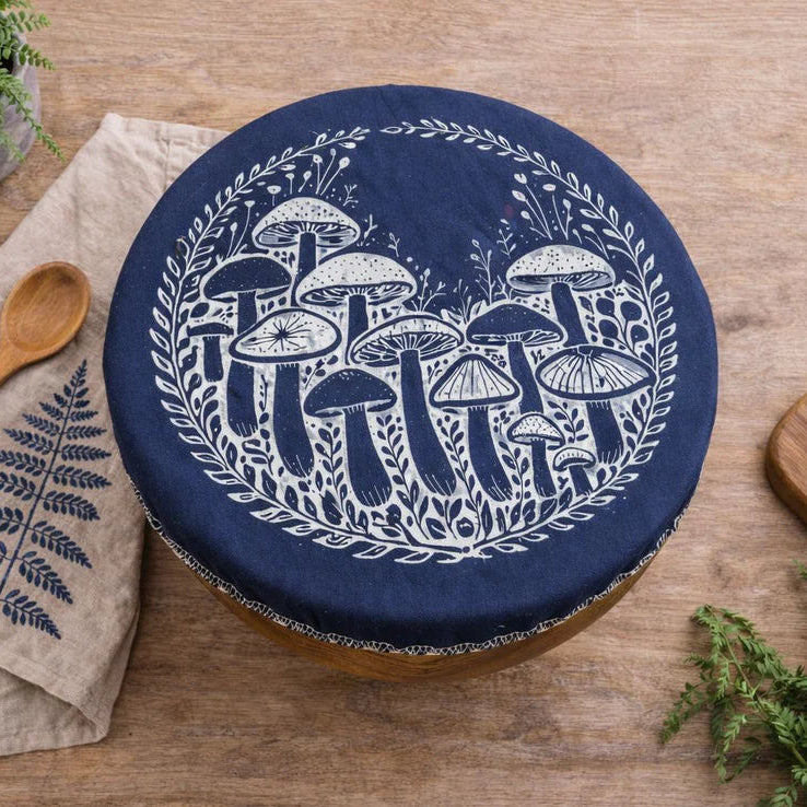 Navy blue fabric bowl cover with white mushroom design on a wooden surface with bread and a plant.