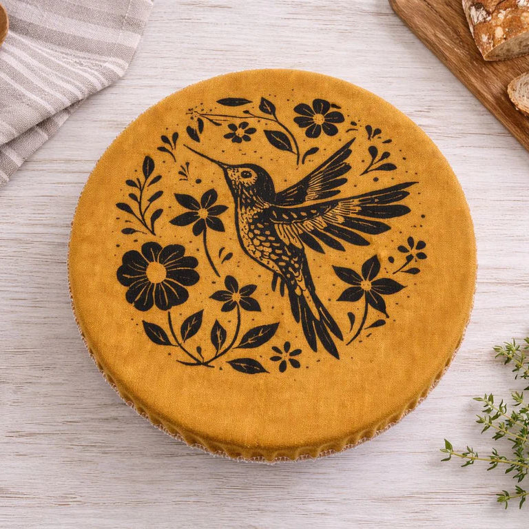 Wooden cutting board with a bowl covered with a golden cloth bowl cover with a hummingbird and floral design.