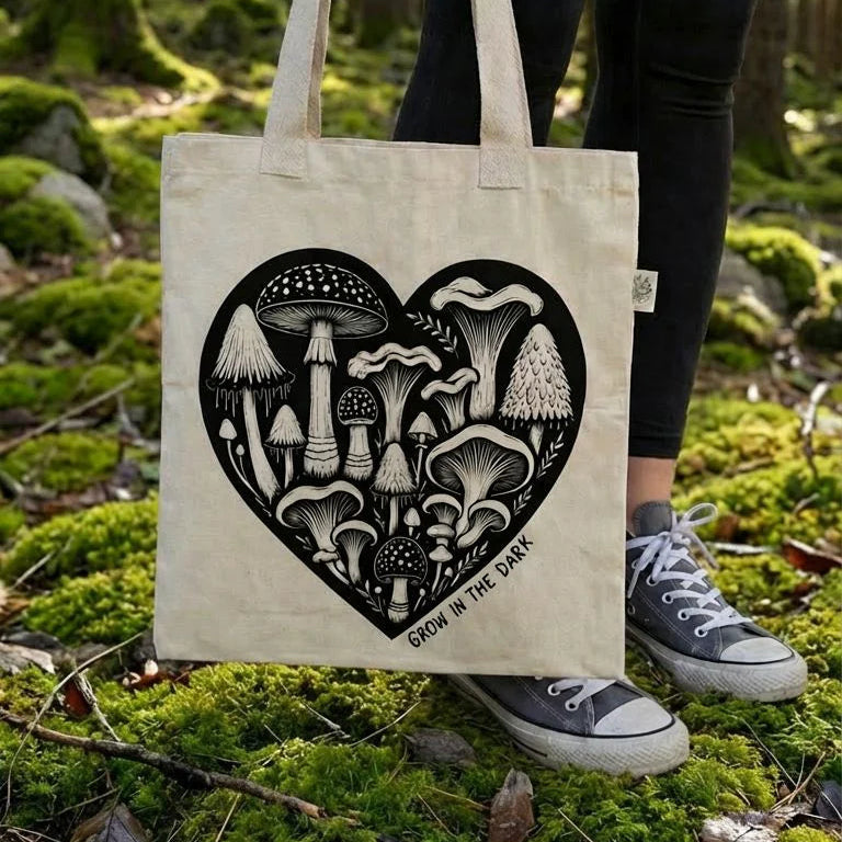 Person holding a tote bag with a heart-shaped mushroom design that says "grow in the dark" in a forest setting.
