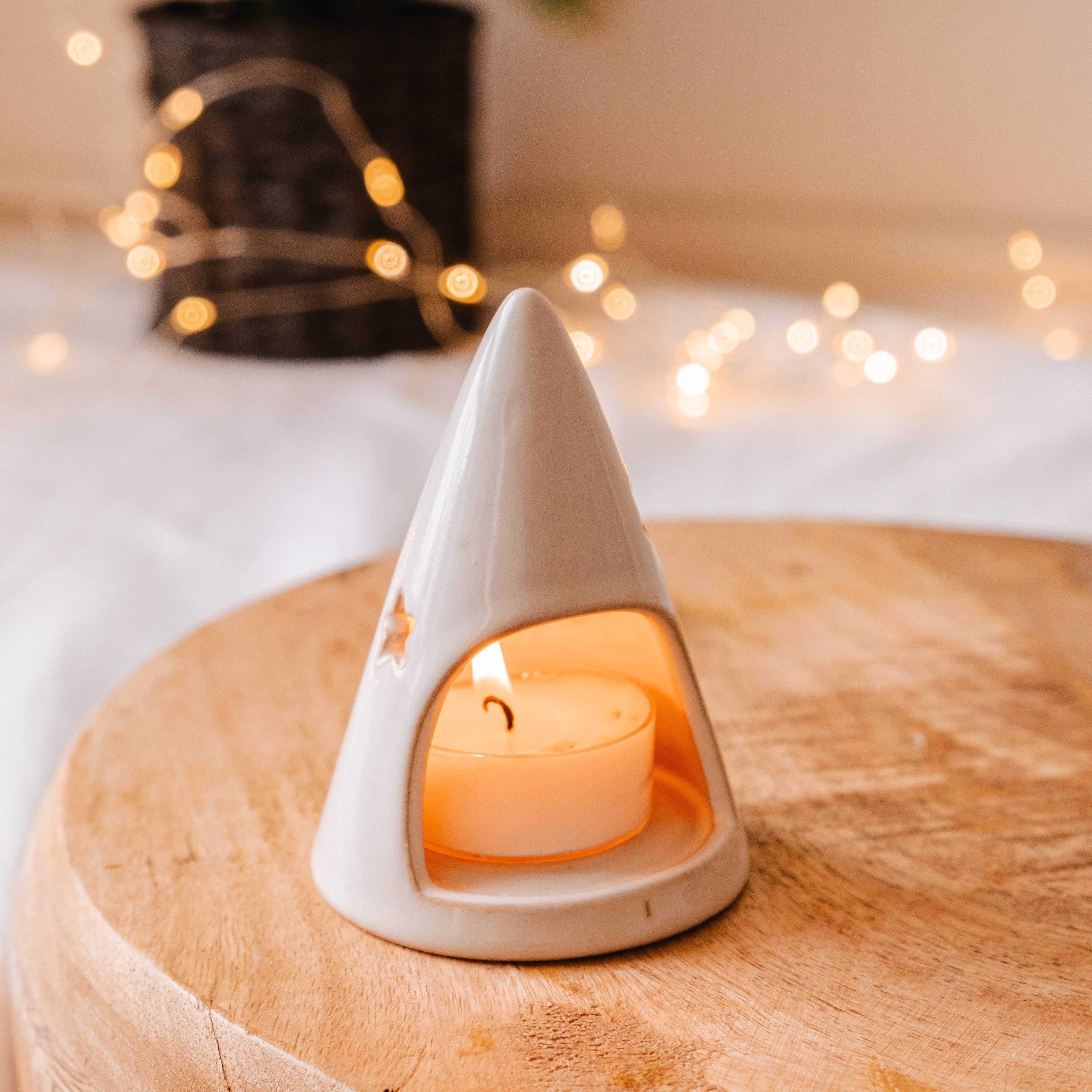 White conical candle holder with a lit candle on a wooden surface with blurred lights in the background