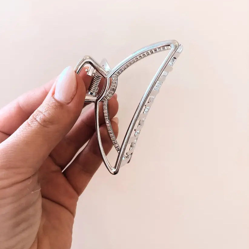 A silver metal claw hair clip held by a hand against a plain light beige background.