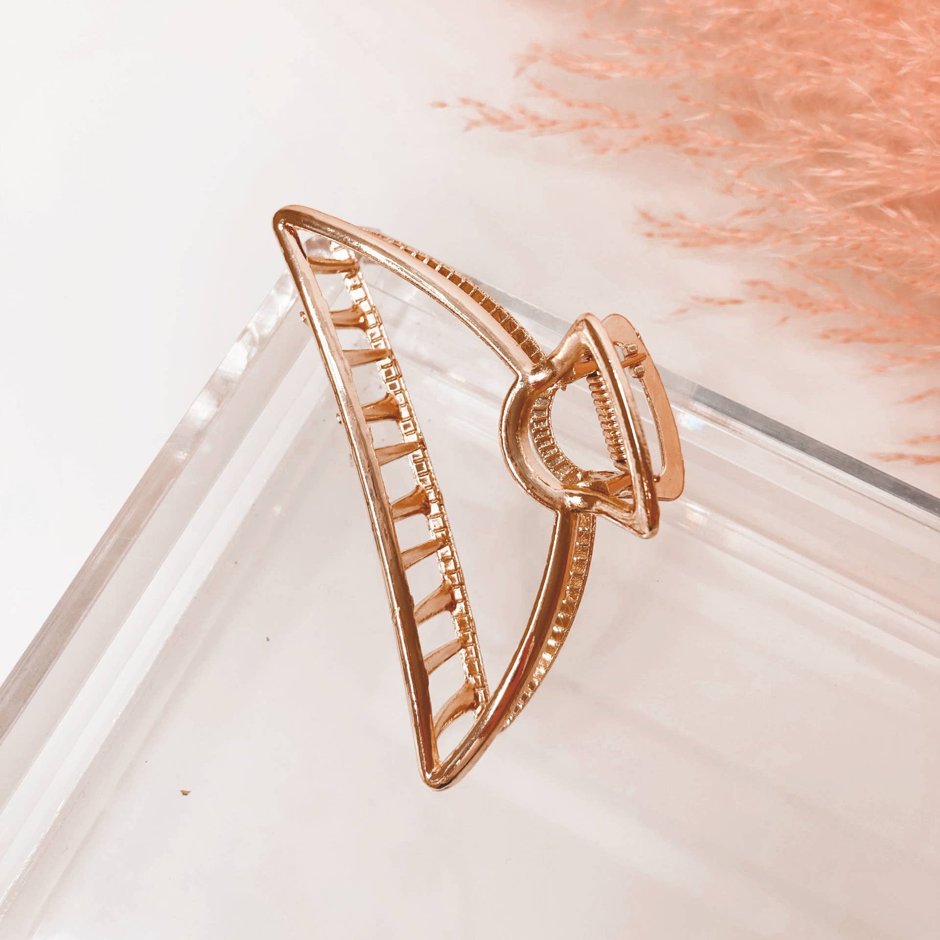 A gold-toned hair claw clip with a textured, open geometric design, resting on a clear acrylic tray with soft peach pampas grass in the background.