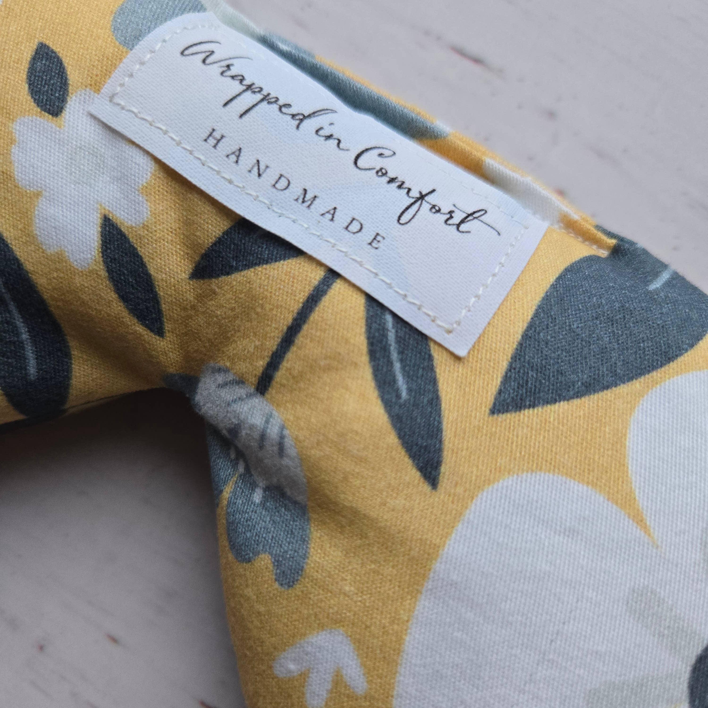organic cotton bean shaped heating pad with floral-patterned fabric with a 'Wrapped in Comfort' label on a wooden surface